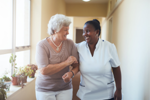 Care worker in uniform walking arm-in-arm with a smiling elderly woman down a care home corridor